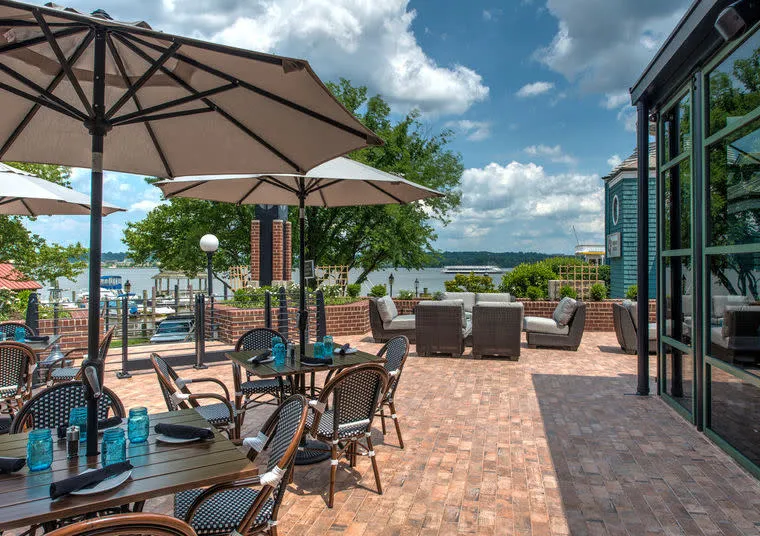 Outdoor restaurant patio with tables, chairs, and large umbrellas on a brick floor, overlooking a river with trees and boats in the background under a partly cloudy sky.