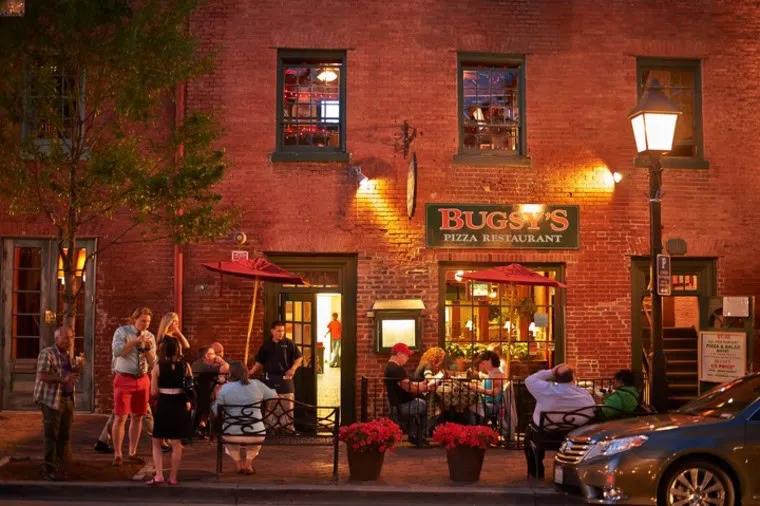 People dine and socialize outside Bugsys Pizza Restaurant in the evening, seated at tables along a brick building with red umbrellas and warm lighting. A tree and streetlamp line the sidewalk, and a car is parked nearby.