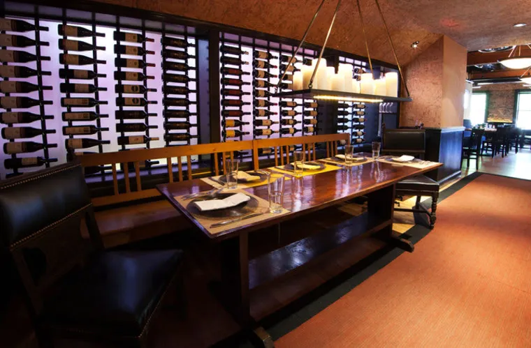 A long wooden dining table is set with plates, napkins, glasses, and cutlery in a warmly lit restaurant. Behind the table is a decorative wine rack filled with bottles. Modern pendant lights hang above.