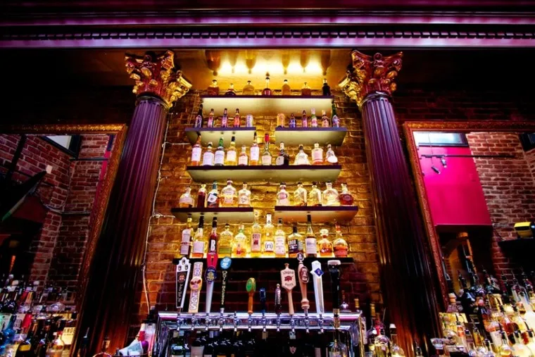 A bar interior with illuminated shelves displaying bottles of liquor between two ornate columns, with beer taps below and brick walls in the background.