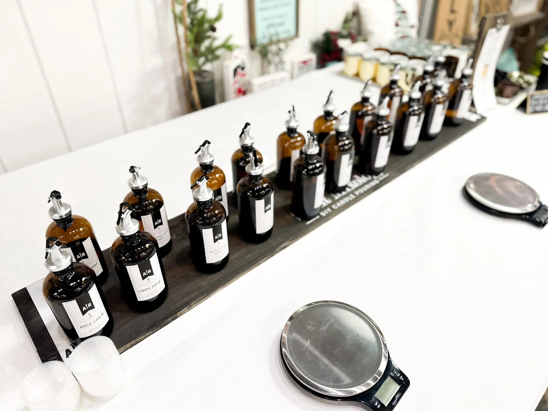 A row of brown glass bottles with pump dispensers and white labels sits on a long black tray atop a white table, flanked by digital scales and small plastic cups; plants and decor appear in the background.