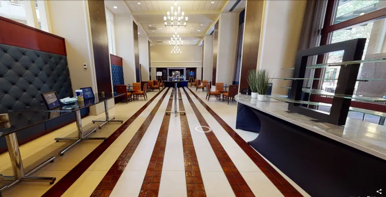 A spacious hotel lobby with striped tile flooring, tall ceilings, chandeliers, and seating areas along the sides. Glass display tables and a plant decorate the right side near large windows.