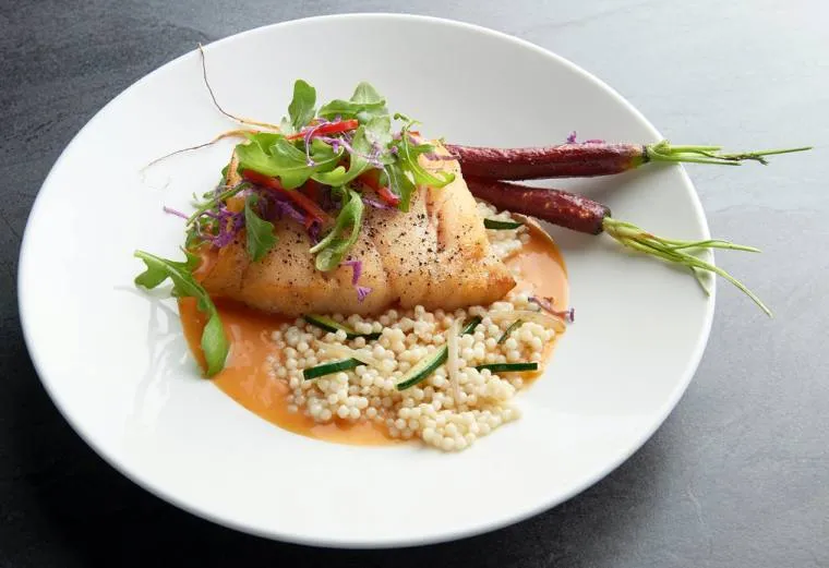 A white plate with a fillet of fish topped with greens, served with sauce, pearl couscous, and two whole glazed carrots with stems on a dark surface.
