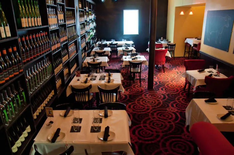 A cozy restaurant with tables set for dining, each covered with white tablecloths and black napkins. Walls are lined with wine bottles, and the red carpet features a bold circular pattern. Soft lighting creates a warm ambiance.