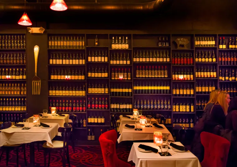 A cozy restaurant with shelves full of wine bottles, dim lighting, red and black decor, and tables set with white tablecloths and candles. Two people are seated and conversing at a table on the right.