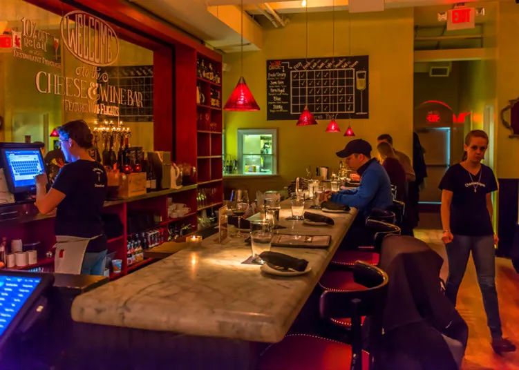 A cozy wine bar with a marble counter, red bar stools, and dim lighting. A bartender works at a register, a few patrons sit at the bar, and a server walks by. Wine bottles and glasses decorate the shelves.