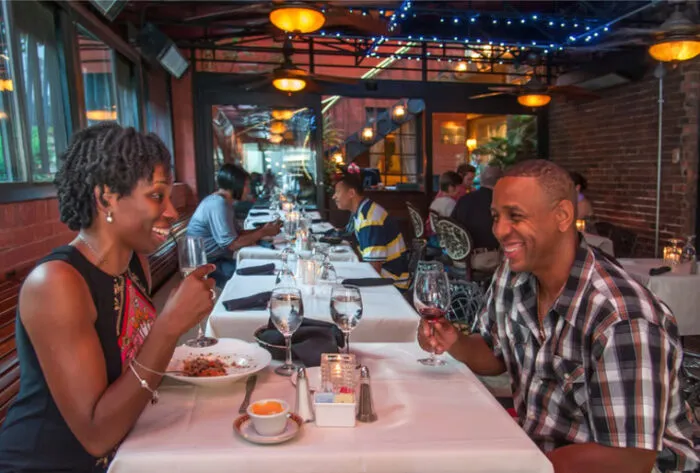 A smiling couple enjoys wine and dinner at a restaurant with brick walls, string lights, and other diners seated at tables in the background.