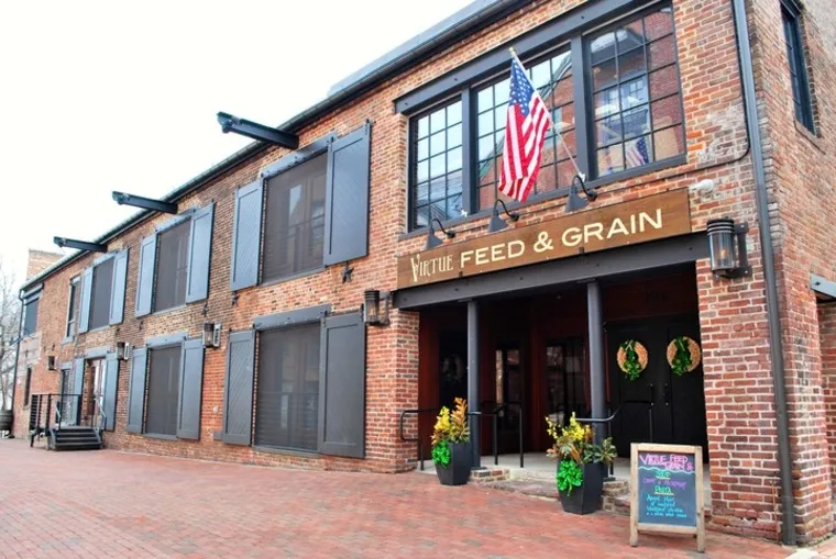 A brick building with black shutters houses Virtue Feed & Grain. An American flag hangs above the entrance, which is decorated with wreaths. A chalkboard sign and potted plants sit outside on the brick sidewalk.