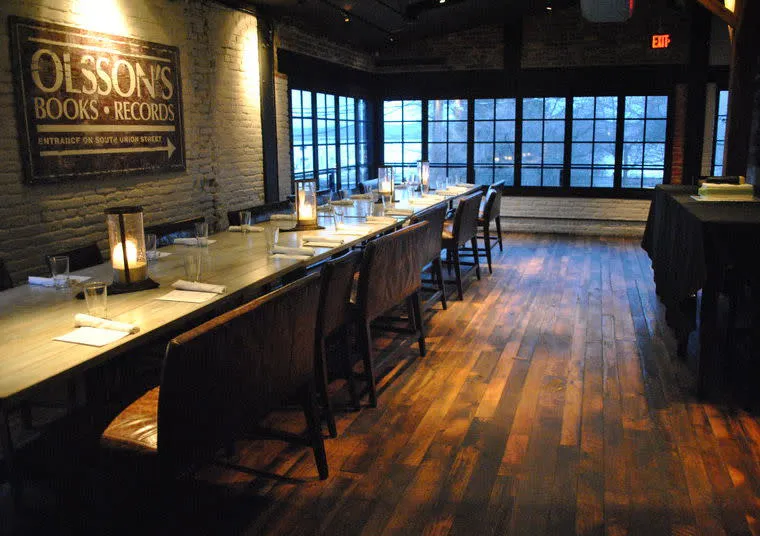 A long dining table with chairs is set for a meal in a cozy, dimly lit room with wooden floors, brick walls, large windows, and a vintage sign reading OLSSONS BOOKS RECORDS.