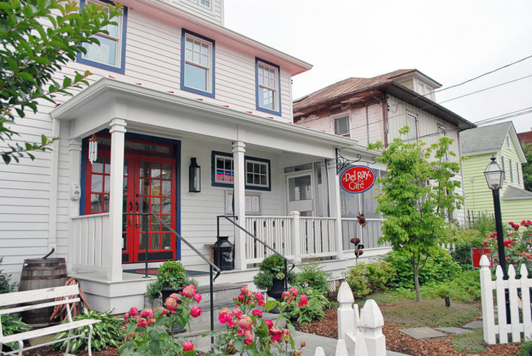 A white, two-story house with a front porch, red door, and a “Del Ray Café” sign hangs by the entrance. Pink flowers and greenery line the yard, bordered by a white picket fence. Neighboring houses are visible.