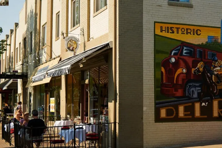 Outdoor dining tables with people sit along a sidewalk outside a restaurant; a large mural with a red train and the words “Historic Del Rio” is painted on the side of a brick building.