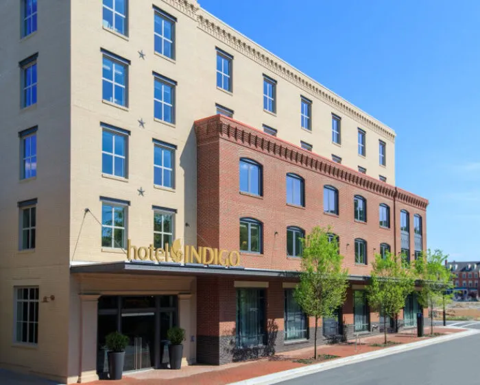 A modern, four-story Hotel Indigo building with beige and red brick exterior, large windows, gold sign, and small trees along the sidewalk on a sunny day.