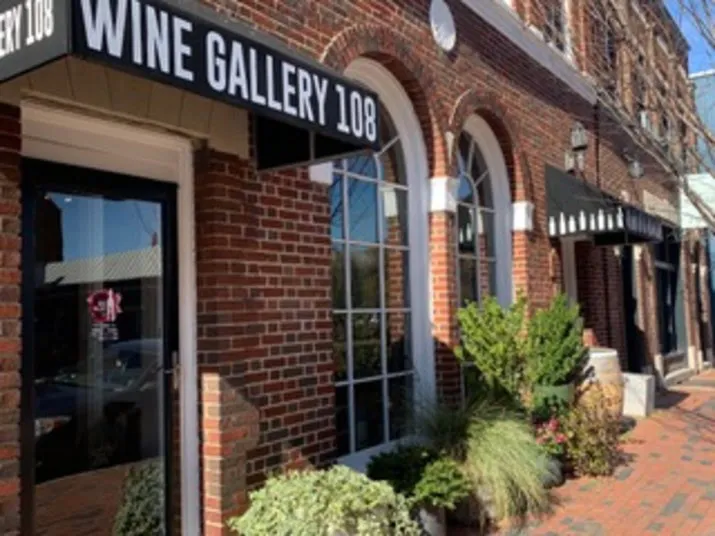 A brick building with large arched windows and a black awning reading WINE GALLERY 108. Potted plants line the sidewalk and nearby storefronts have matching awnings.