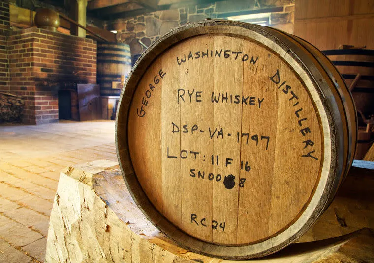 A wooden whiskey barrel labeled George Washington Distillery Rye Whiskey sits in a rustic distillery with stone walls and brick fireplace in the background, illuminated by warm natural light.