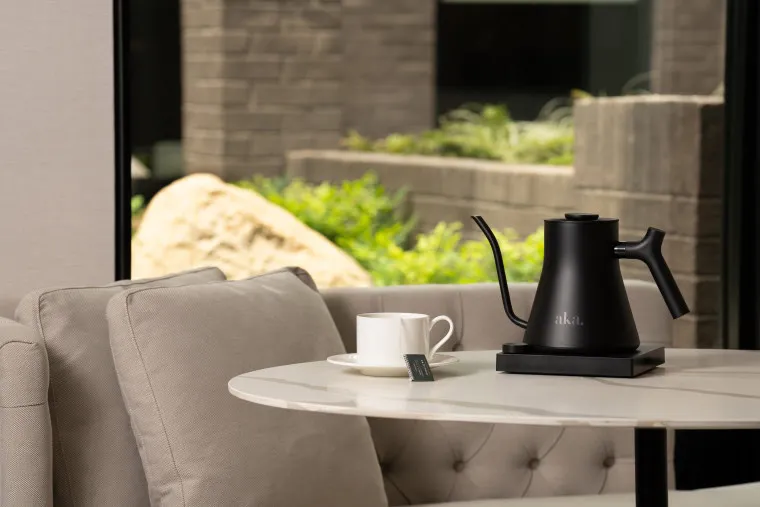 A modern black electric kettle and a white teacup with a tea bag sit on a round marble table next to a light gray couch, with a view of greenery and stone through a large window in the background.