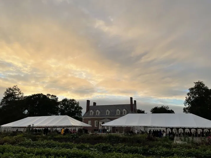Two large white tents are set up on a lawn in front of a historic brick house at sunset, with groups of people gathered beneath the tents and a dramatic, cloudy sky overhead.