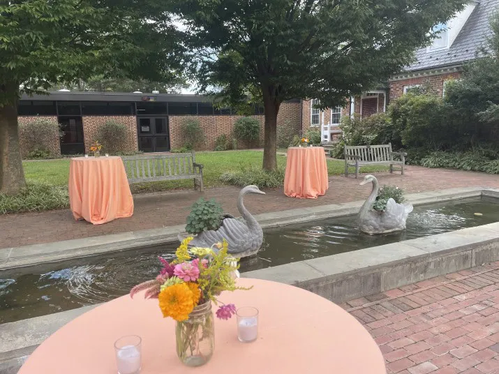 A courtyard with brick paving features a small fountain with two swan sculptures. Tables covered in peach cloths, floral centerpieces, and benches are arranged under leafy trees beside a brick building.