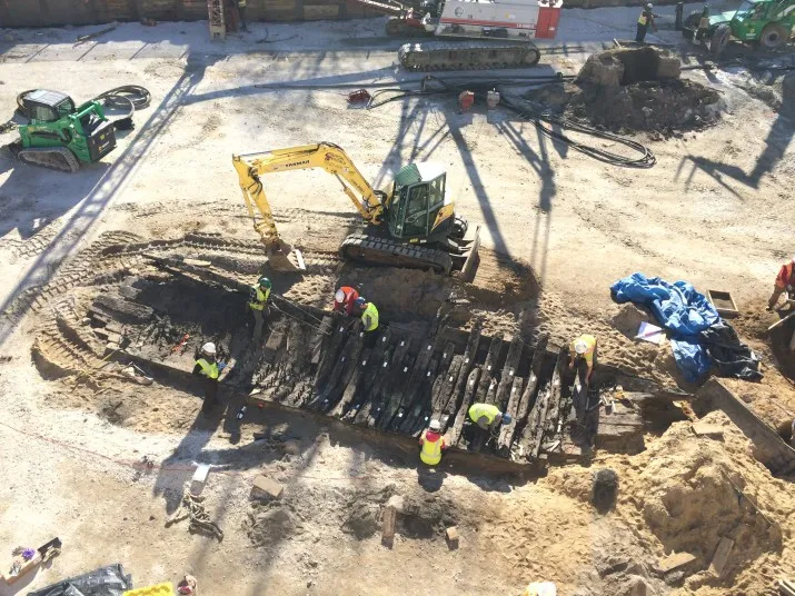A group of workers in safety vests excavate and study the wooden remains of an old ship buried at a construction site, with construction vehicles and equipment surrounding the area.