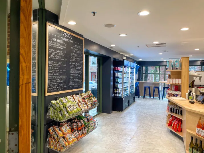 A small market interior with shelves of chips, a chalkboard menu on the wall, refrigerated drink cases, snack displays, and a counter area with bar stools near large windows.