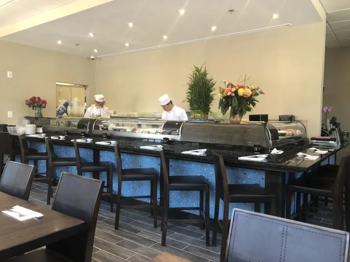 Two sushi chefs work behind a counter in a modern Japanese restaurant. Fresh flowers and plants decorate the counter, while neatly arranged tables and chairs fill the dining area.