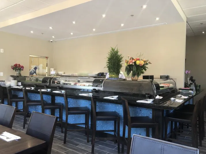 A modern sushi bar with a blue-tiled counter, dark chairs, floral arrangements, and sushi displayed in glass cases. The setting is bright with overhead lights and neatly arranged utensils on the tables.