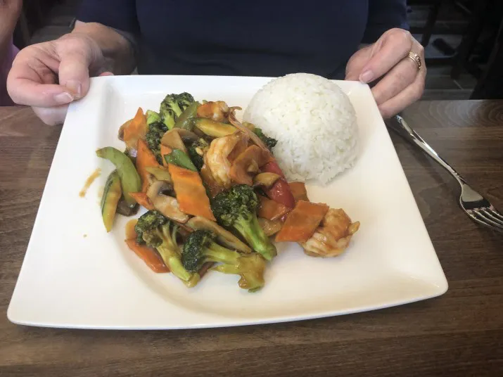 A person holds a square white plate with steamed white rice and stir-fried vegetables including broccoli, carrots, and zucchini in sauce, served on a wooden table.