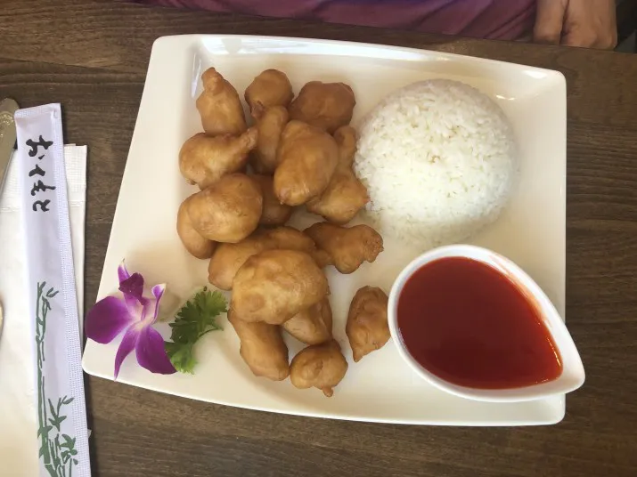A square white plate with battered fried chicken pieces, a mound of white rice, a small bowl of red sweet and sour sauce, a sprig of parsley, and a purple orchid flower garnish. Chopsticks are placed to the side.