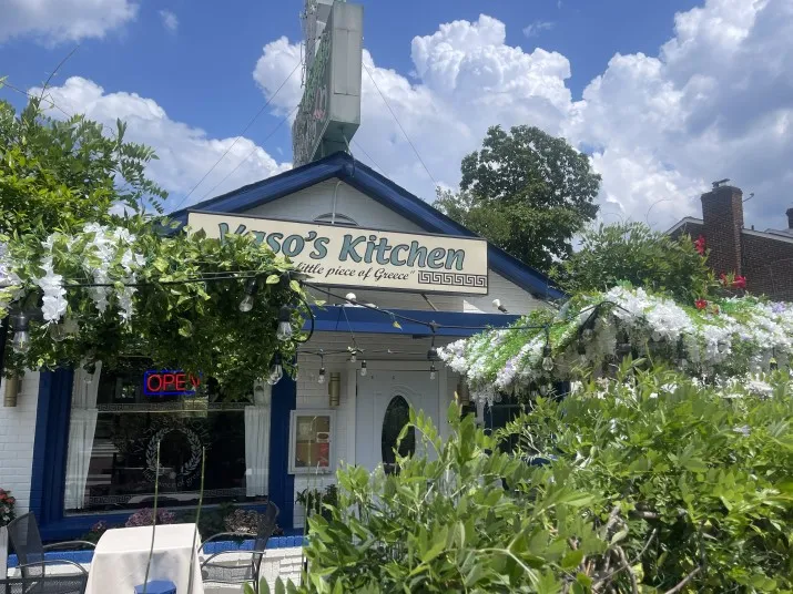 A white and blue restaurant called Vasos Kitchen with a sign reading A little piece of Greece. The entrance is surrounded by greenery, flowers, and outdoor seating. A neon OPEN sign is visible under a partly cloudy sky.