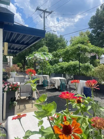 Outdoor patio of a restaurant with tables and chairs set under umbrellas, surrounded by colorful red flowers and green plants, beneath a bright blue sky with scattered clouds.