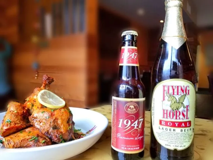 A plate of roasted chicken garnished with a lemon slice sits next to two bottles of beer labeled 1947 and Flying Horse Royal on a wooden table in a warmly lit restaurant.