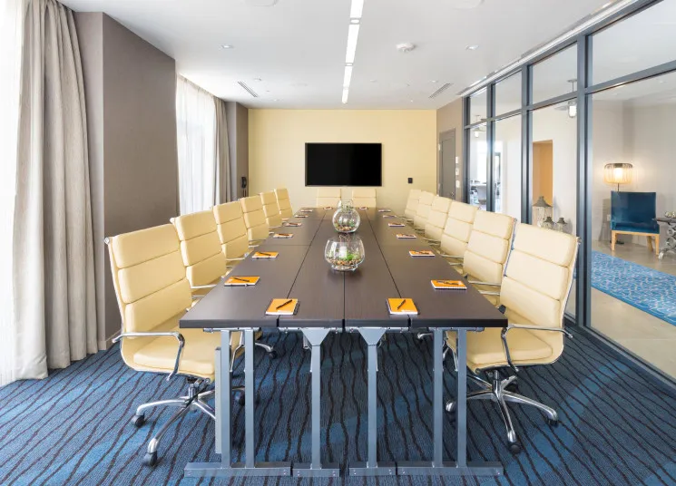 Modern conference room with a long table, beige swivel chairs, notepads and pens, glass vases as centerpieces, large screen on the far wall, floor-to-ceiling windows, and blue carpet flooring.