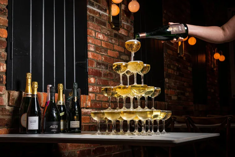 A person pours champagne into a pyramid of stacked glasses on a table, with several bottles of champagne next to the tower against a brick wall and warm lighting.