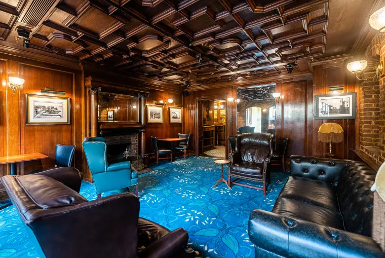 A luxurious, wood-paneled lounge features leather armchairs, framed black-and-white photos, a fireplace, vintage lamps, and a blue carpet with a floral pattern. The coffered ceiling adds to the classic, elegant atmosphere.