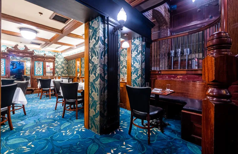 Elegant restaurant interior with floral blue carpet, dark wood furniture, leafy wallpaper, and classic light fixtures. Tables are set with white linens and a vintage wood partition divides the room.