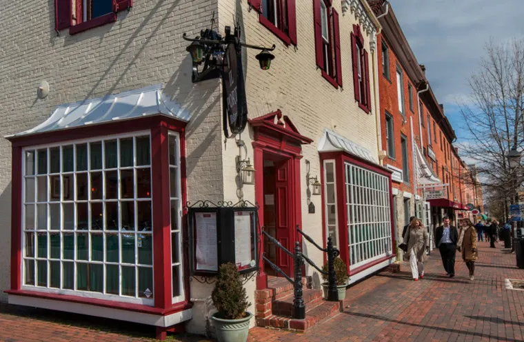 A street scene with people walking past a cream-colored building featuring large, grid-pane windows and red trim, located along a brick sidewalk on a partly cloudy day.