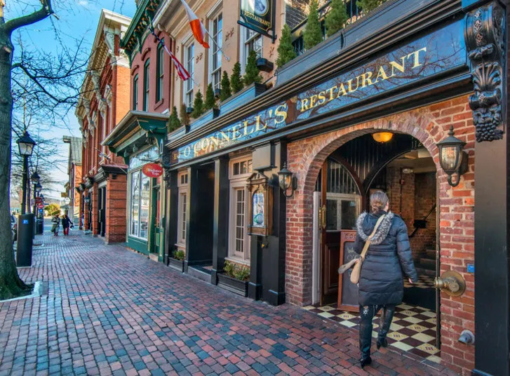 A woman in a black coat exits O’Connell’s Restaurant, located in a historic brick building along a charming cobblestone street lined with shops and old-fashioned street lamps.