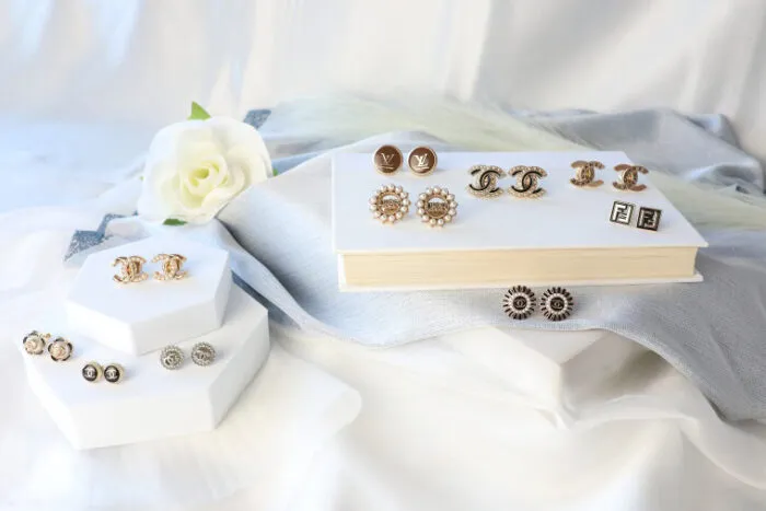 Gold and silver cufflinks with various designs, including logos and floral shapes, are displayed on white hexagonal stands and a book, with a white rose and light fabric in the background.