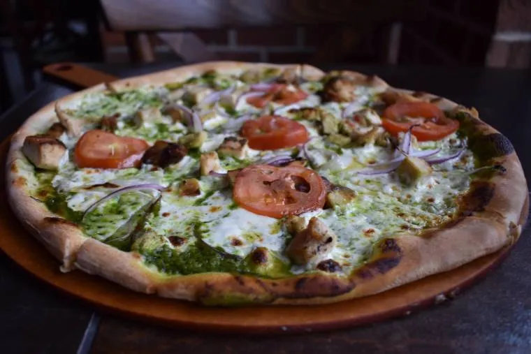 A freshly baked pizza topped with sliced tomatoes, red onions, chicken pieces, mozzarella cheese, and green pesto sauce sits on a wooden serving board on a dark table.
