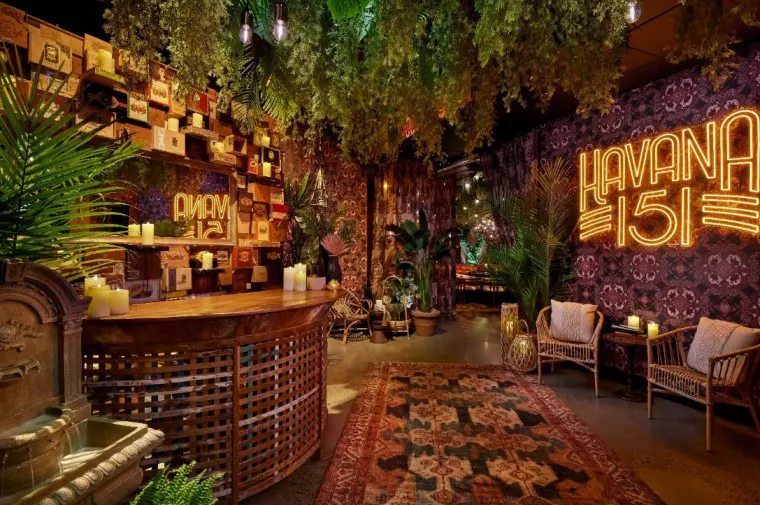 A cozy, Havana-themed lounge with lush plants, vintage decor, patterned rugs, wicker chairs, a wooden bar, and warm, glowing “Havana 151” neon sign on a wallpapered wall. Candles and books add to the inviting atmosphere.