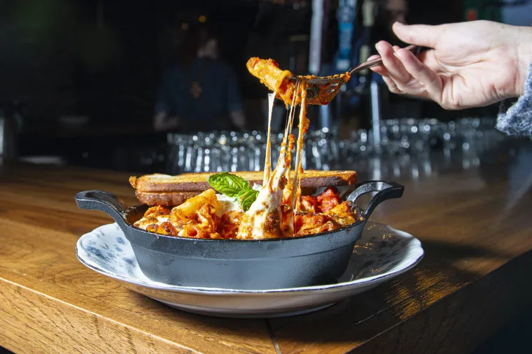 A hand lifts a cheesy pasta bake with a fork from a black cast iron dish, revealing melted cheese stretching. The dish sits on a white plate on a wooden table, with a breadstick and basil garnish on top.
