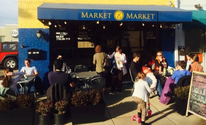 People enjoying food and drinks at outdoor tables in front of a café called Market Market on a sunny day. Children play nearby while adults chat and dine under a blue awning.