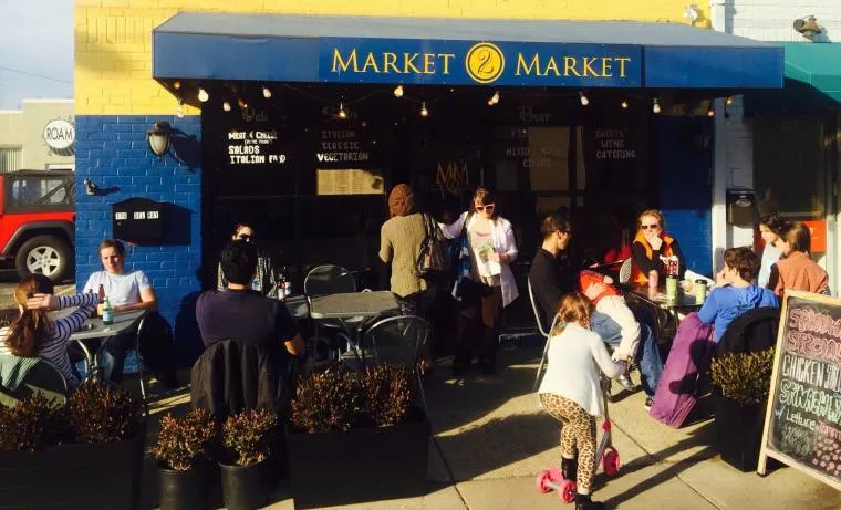 People enjoying food and drinks at outdoor tables in front of a café called Market Market on a sunny day. Children play nearby while adults chat and dine under a blue awning.