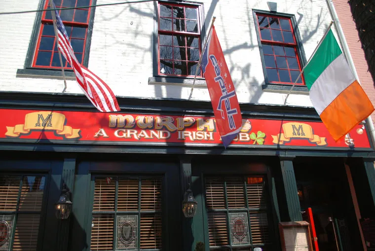 The exterior of an Irish pub with a red sign reading Murphys A Grand Irish Pub, flanked by American, Irish, and another flag, with green trim and large windows beneath the sign.