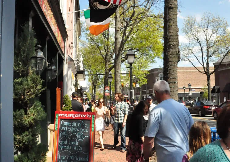 People walk along a busy sidewalk outside Murphy’s pub on a sunny day. A chalkboard menu stands by the entrance, and American and Irish flags hang above. Trees with green leaves line the street.