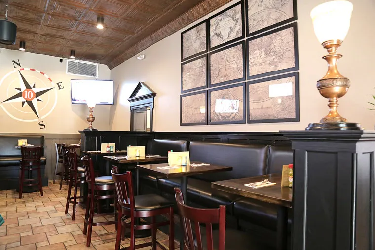 A cozy restaurant interior with dark wooden booths and chairs, maps on the wall, a compass mural, and a lit lamp on a black partition. A TV is mounted on the wall above the seating area.