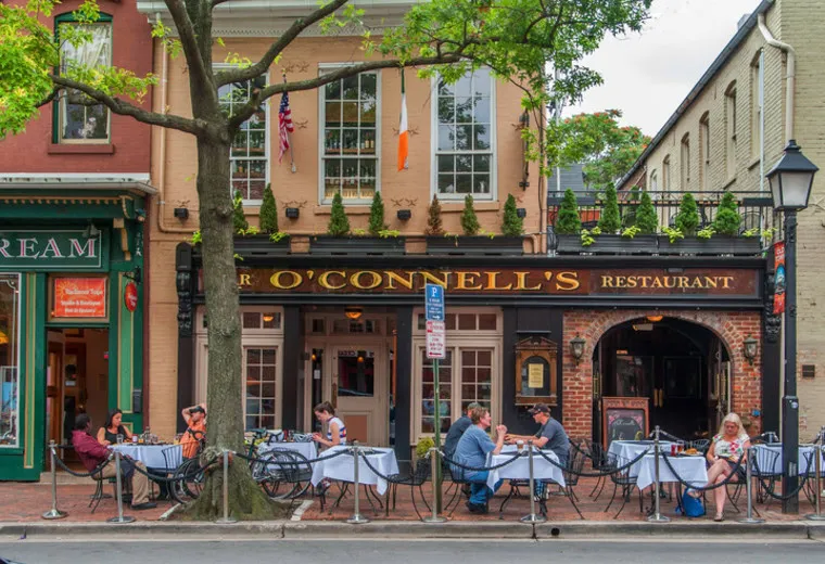 People dine outdoors at tables with white tablecloths in front of an Irish restaurant called O’Connell’s. The scene is lively with trees, flags, and neighboring buildings visible on a sunny day.