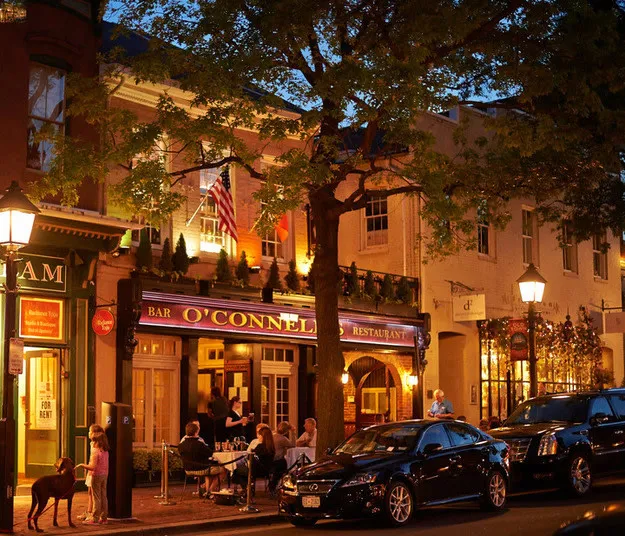 People dine outdoors at a lively restaurant called O’Connell’s, illuminated by warm lights at dusk. Cars are parked along the street and a dog stands near a lamppost under leafy trees.