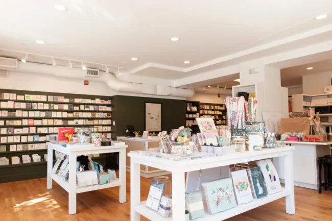 A bright, modern stationery store with white tables displaying greeting cards, notebooks, and art prints. Shelves along the back wall are filled with colorful cards. Wood floors and white walls create an inviting, organized space.