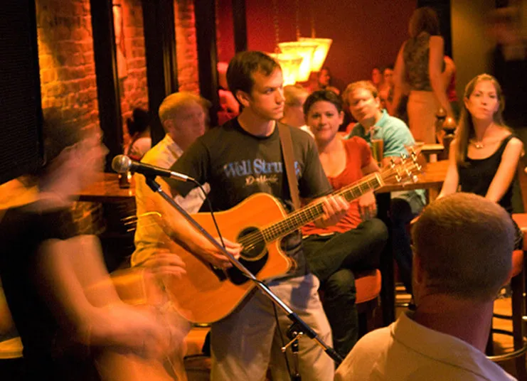 A man plays an acoustic guitar and sings into a microphone in a cozy, dimly lit venue, surrounded by people sitting and watching attentively. Warm lights and relaxed atmosphere suggest a casual live music event.