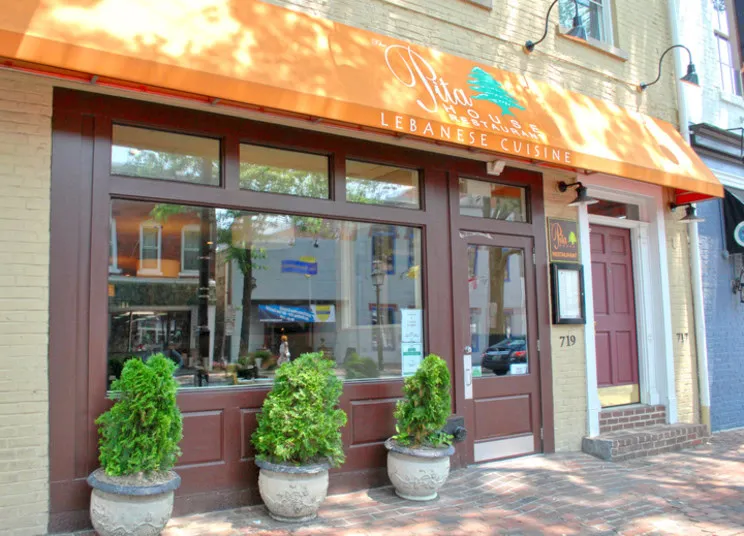 A storefront with an orange awning that reads The Pita House Lebanese Cuisine. The entrance is flanked by potted plants, and large windows reflect trees and buildings across the street. The address number “719” is visible.